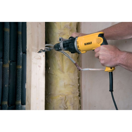 A person using a yellow DEWALT electric drill to bore a hole through a wooden beam, with electrical wires passing through the opening in a construction or renovation setting.