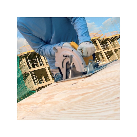 A person wearing gloves uses a DEWALT circular saw to cut a sheet of plywood at a construction site. The building structure and scaffolding are visible in the background.