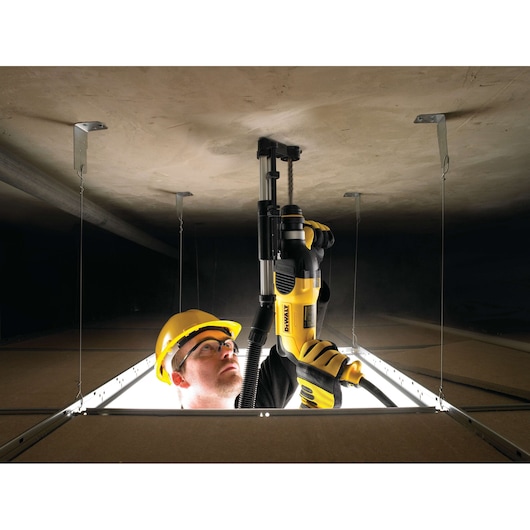 A person wearing a yellow safety helmet and gloves is using a DEWALT D25301D power tool to drill into a ceiling, seen from below through an open ceiling panel. The face of the person is blurred for privacy.