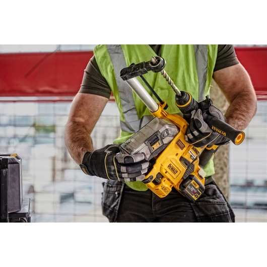 A person wearing a green safety vest and work gloves is holding a yellow DEWALT cordless rotary hammer drill with a dust extraction attachment, in a construction site setting.