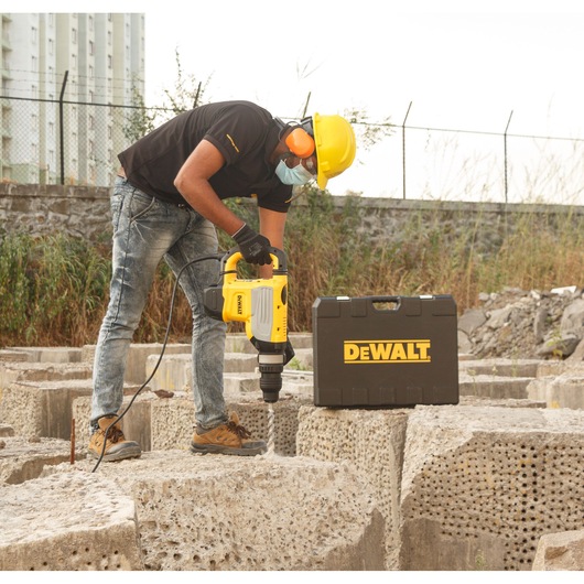 A person wearing safety gear, including a helmet, gloves, ear protection, and a mask, uses a DEWALT hammer drill on a large concrete block. A DEWALT branded tool case is placed nearby.