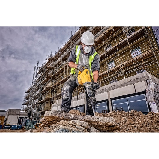 A construction worker wearing a high-visibility vest and a white hard hat uses a yellow DEWALT demolition hammer drill to break up concrete at a construction site with scaffolding and a building under construction in the background.