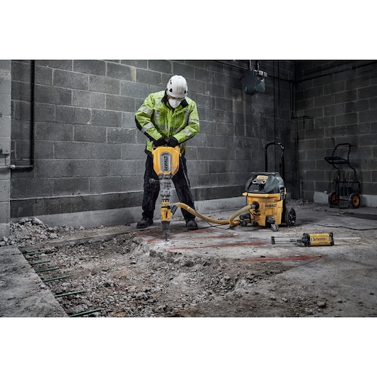 A construction worker in safety gear operates a DEWALT demolition hammer (model D25911) to break up concrete on a worksite. The worker is wearing a helmet and high-visibility jacket. A DEWALT vacuum extractor is connected to the tool, and other equipment is visible in the background.