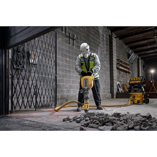 A worker wearing safety gear operates a yellow DEWALT jackhammer, breaking concrete in an industrial setting. Broken concrete pieces are scattered on the ground, and DEWALT equipment is visible in the background.