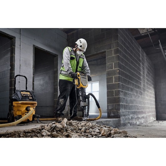 A construction worker wearing a safety vest and helmet is using a DEWALT jackhammer (model D25966) to break up concrete inside an unfinished building. There is a DEWALT dust extractor connected to the jackhammer, and broken concrete rubble is scattered on the floor.