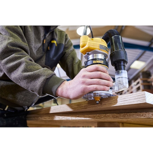 A person using a DEWALT D26200 router tool with a dust extraction attachment on a piece of wood in a workshop.
