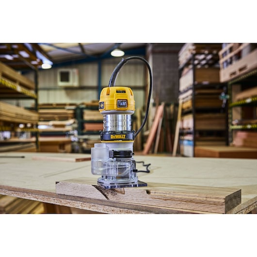 A DEWALT D26200 compact router placed on a wooden board in a workshop, surrounded by shelves and stacks of wood.