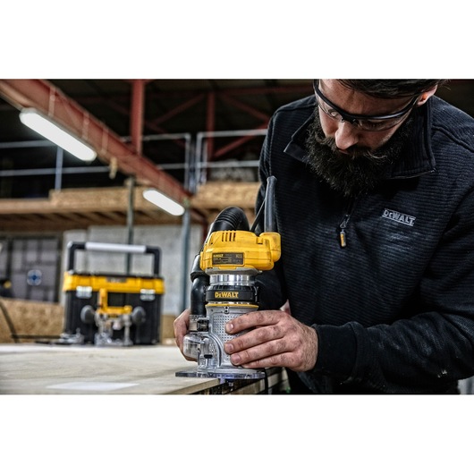 A person using a DEWALT D26204K power tool to rout the edge of a wooden board in a workshop. The individual is wearing a dark DEWALT-branded jacket, and another DEWALT tool is visible in the background.