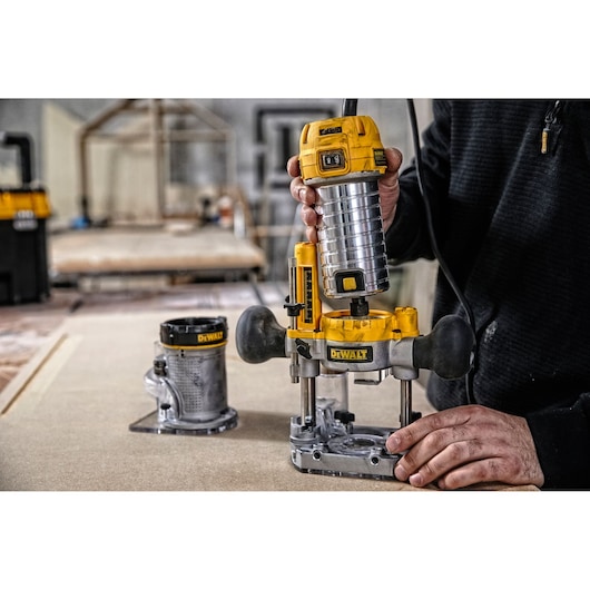 A person using a DEWALT D26204K woodworking router on a workbench, with an additional router attachment placed nearby. The scene is set in a workshop environment.