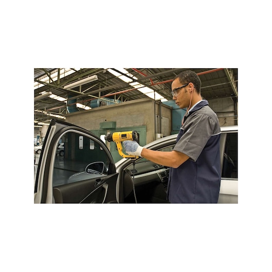 A person wearing gloves is using a yellow DEWALT heat gun (model D26411) on the interior of a car door in a workshop setting. The face of the person is blurred for privacy.