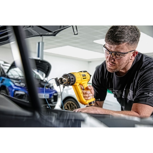 A person using a yellow DEWALT heat gun (model D26414) inside a car workshop. The person's face is blurred, and there are cars with open hoods in the background.