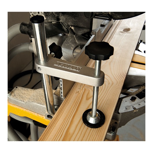 A close-up view of a DEWALT mitre saw with a wood board clamped securely using a metal clamp. The saw is set up for precise woodworking with adjustment knobs and a dust extraction hose visible in the background.
