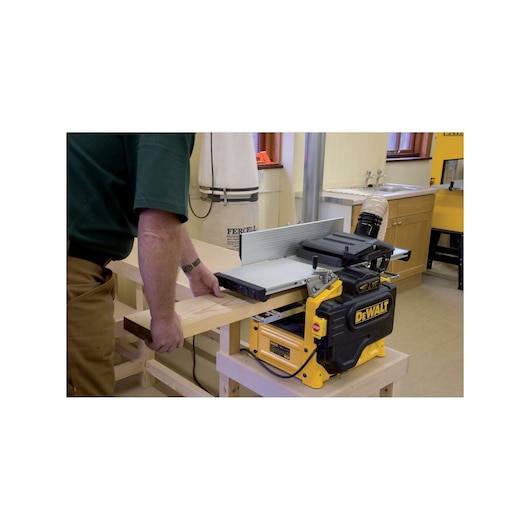 A person is operating a DEWALT woodworking planer (model D27300) in a workshop, processing a wooden board. The planer is placed on a sturdy bench and connected to a dust extraction system.