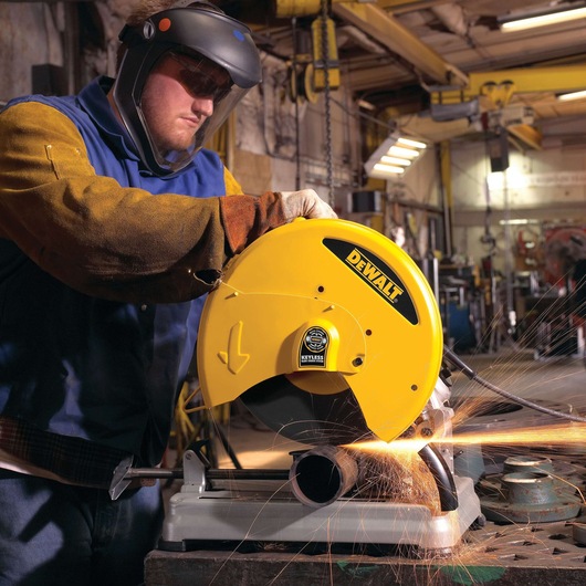 A worker wearing protective gear operates a DEWALT D28715 metal chop saw, cutting a metal pipe and producing sparks in an industrial workshop setting.