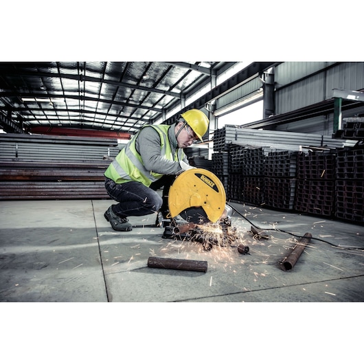 A worker wearing a safety vest and helmet uses a DEWALT D28730-B2 metal chop saw to cut steel pipes in an industrial warehouse. Sparks fly from the cutting area and there are steel pipes and stacked metal materials in the background.