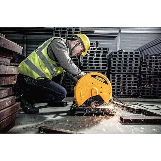 A person wearing safety gear operates a DEWALT metal cutting saw, model D28730, to cut metal bars in an industrial setting. Sparks fly from the cutting area.