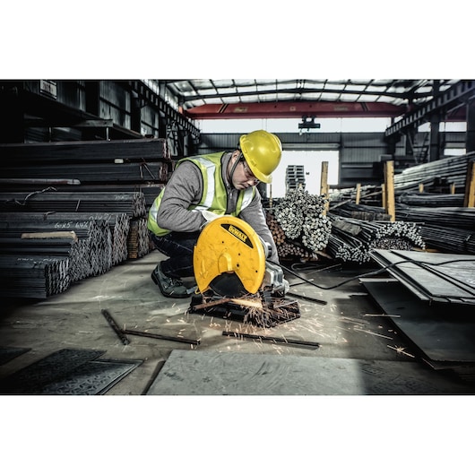 A worker wearing a yellow safety helmet and reflective vest is cutting metal with a yellow DEWALT chop saw, producing sparks. The worker is in an industrial warehouse surrounded by stacks of metal bars and sheets.