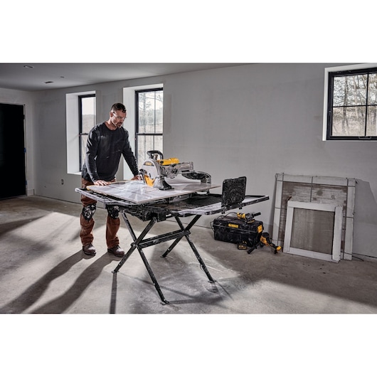 A person is working indoors with a DEWALT D36000 tile saw placed on a folding stand. The workspace appears to be a construction or renovation site, with tools and materials nearby, including a DEWALT tool bag and a window screen leaning against the wall.
