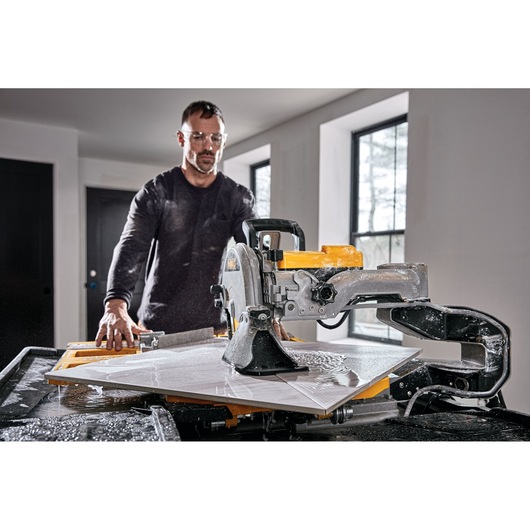 A person operates a DEWALT D36000 wet tile saw on a worktable, cutting a large white tile inside a modern room. The saw is actively spraying water to cool the blade during the cutting process.