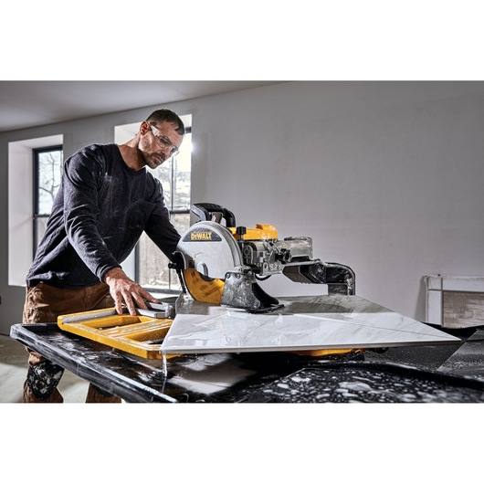 A person operates a DEWALT D36000 tile saw indoors, cutting a large piece of tile on a worktable. The individual's face is blurred for privacy.
