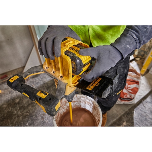 A person wearing gloves uses a yellow DEWALT cordless mixer with a DEWALT DCB548 battery to mix material in a bucket on a construction site.