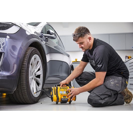 A person kneeling on the floor in a garage is using a DEWALT DCC018N portable inflator to fill the tire of a parked car. The inflator is connected to the tire and the person is operating the device.