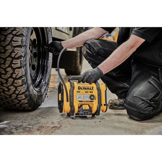 A person wearing black gloves operates a yellow DEWALT portable air compressor to inflate the tire of a vehicle. The air compressor is placed on a concrete floor and connected to the tire with a hose.