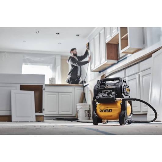 A cordless DEWALT air compressor is placed on the floor of a kitchen being renovated. Cabinets and doors are open or being installed, and a person is working on the upper cabinets in the background.