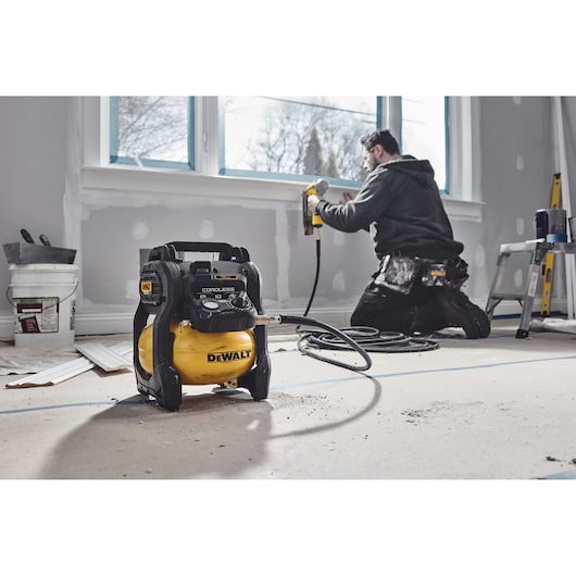 A DEWALT DCC1018N cordless air compressor is placed on the floor of a room under renovation, with a worker using a pneumatic tool in the background near a window.