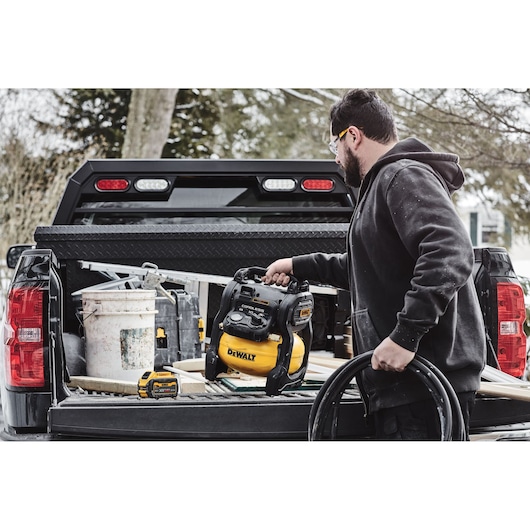 A person places a DEWALT portable air compressor (model DCC1018N) in the back of a pickup truck. The truck bed is filled with construction tools and materials.