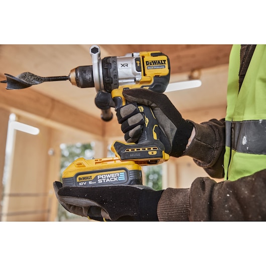 Gloved hands holding a DEWALT cordless drill with POWERSTACK battery, indoors on a construction site.