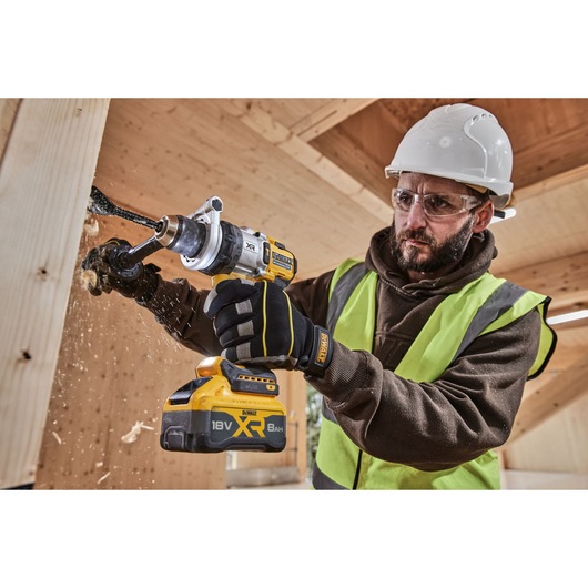 Worker drills wood with DEWALT 18V XR tool, wearing gloves, helmet, and safety vest on construction site.
