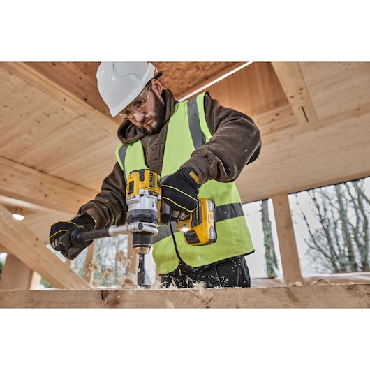 Person uses DEWALT cordless drill to bore wood, wearing safety gear and gloves at a wooden construction site.
