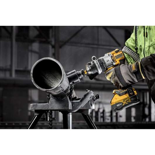 Person using a DEWALT cordless drill to work on a metal pipe inside an industrial setting.