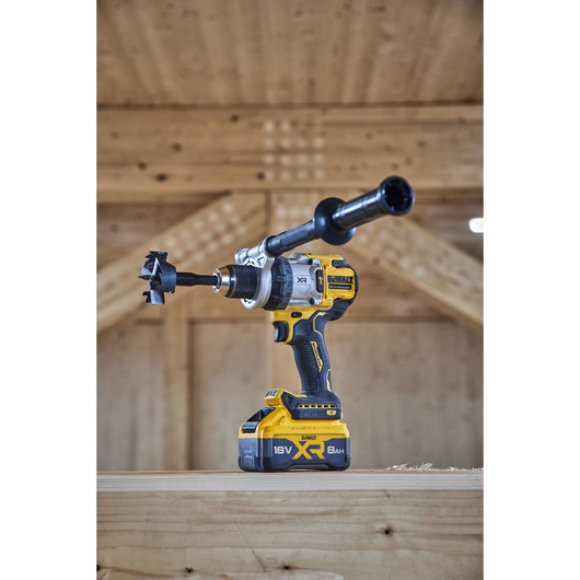A DEWALT 18V XR cordless drill with an auger bit and side handle is placed on a wooden surface in a workshop setting.