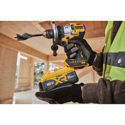 Gloved hand holding a DEWALT 18V XR cordless drill with 8Ah battery in a wooden construction area.