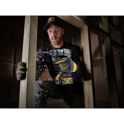 DEWALT DCD100M1T drill used by a worker drilling wood in a construction frame, sawdust flying, gloves worn.