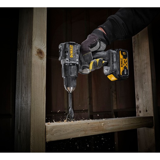 Gloved hand using a DEWALT cordless drill to bore a hole into a wooden beam, wood chips flying.