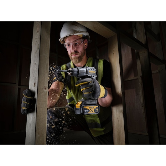 Worker drills wood frame using DEWALT DCD100M2T 18V cordless drill, wearing gloves and safety gear indoors.