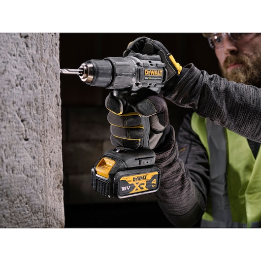 Hand holding a DEWALT cordless drill, drilling into concrete. The drill has an 18V XR 4Ah battery.