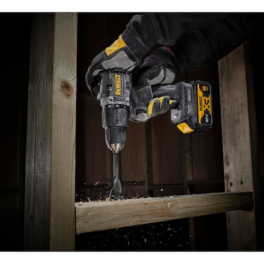 A person wearing protective gloves uses a DEWALT cordless drill to bore a hole in a wooden beam, with wood shavings being produced.