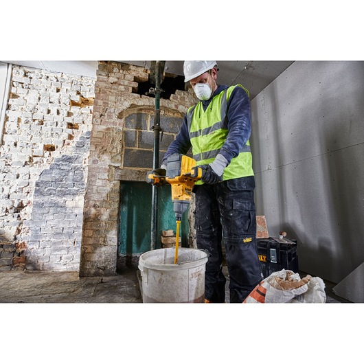 A person wearing a safety vest and gloves is using a DEWALT DCD240X2 cordless mixer to stir contents in a large bucket at a construction site with brick and drywall walls.