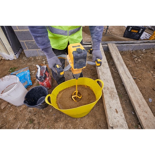 A construction worker is using a yellow DEWALT mixing drill to mix concrete or mortar in a large yellow bucket outdoors. The worker is wearing gloves and a high-visibility vest, with other buckets and bags of materials nearby on the ground.