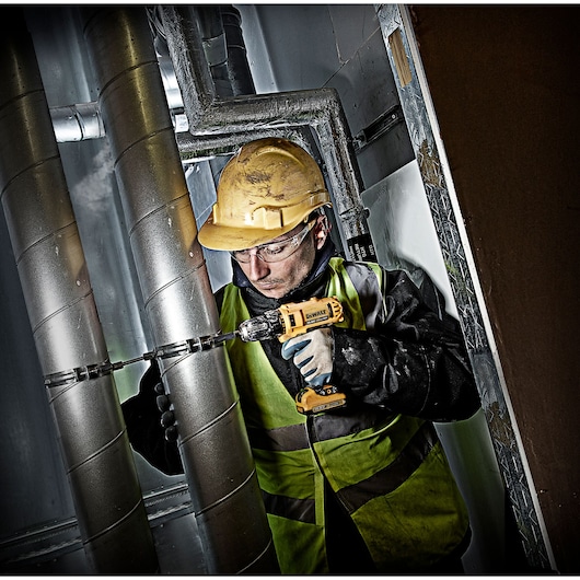 Worker in safety gear uses a DEWALT drill on metal pipes in an industrial setting.