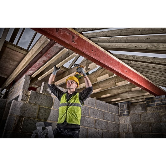 A construction worker wearing a high-visibility vest and hard hat uses a DEWALT cordless drill to secure a wooden beam in the ceiling structure of a building under construction. The worker stands on a ladder surrounded by concrete block walls and timber roof trusses.