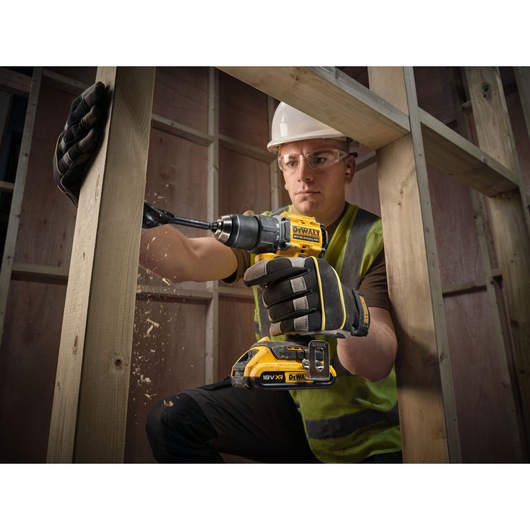 A construction worker wearing gloves and a high-visibility vest is using a DEWALT 18V XR cordless drill to drill into a wooden beam. Sawdust is visible as the drill bit penetrates the wood. The worker's face is blurred and the setting appears to be an unfinished wooden framework.