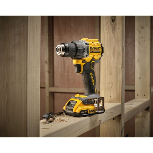 A yellow and black DEWALT cordless drill, model DCD794D2T, is placed upright on a wooden beam in a construction setting. Wood shavings and a drill bit are visible next to the tool.