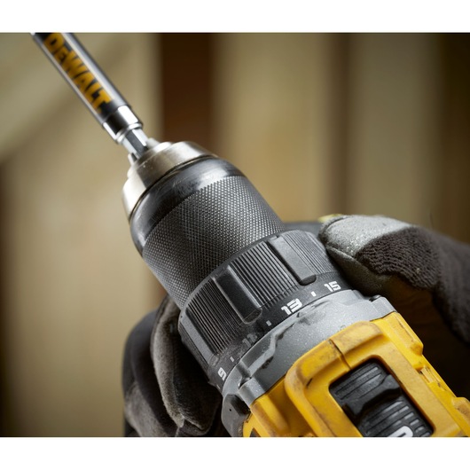 Close-up view of a yellow and black DEWALT cordless drill being held by a gloved hand, showing the adjuster ring and drill bit attached to the chuck.