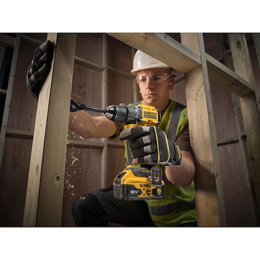 A person wearing protective gloves, a high-visibility vest, and a hard hat is using a DEWALT 18V XR cordless power drill to drill into a wooden frame, with wood shavings visible in the air.