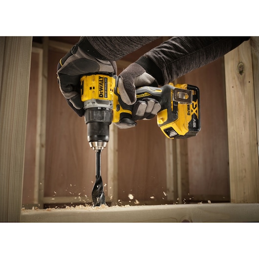 A person wearing gloves is using a DEWALT cordless power drill with a large drill bit to drill into a wooden surface, creating wood shavings. The drill is yellow and black and battery powered.